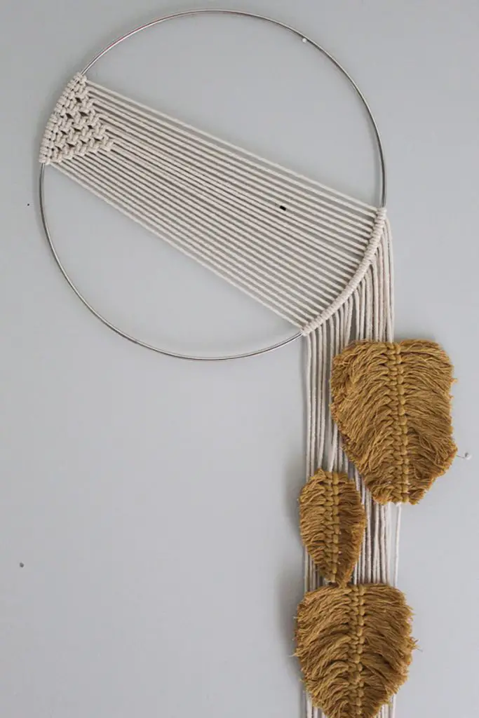 Simple macrame wall hanging with feathers in a mustard color hanging from strings hanging down the right size of the silver hoop